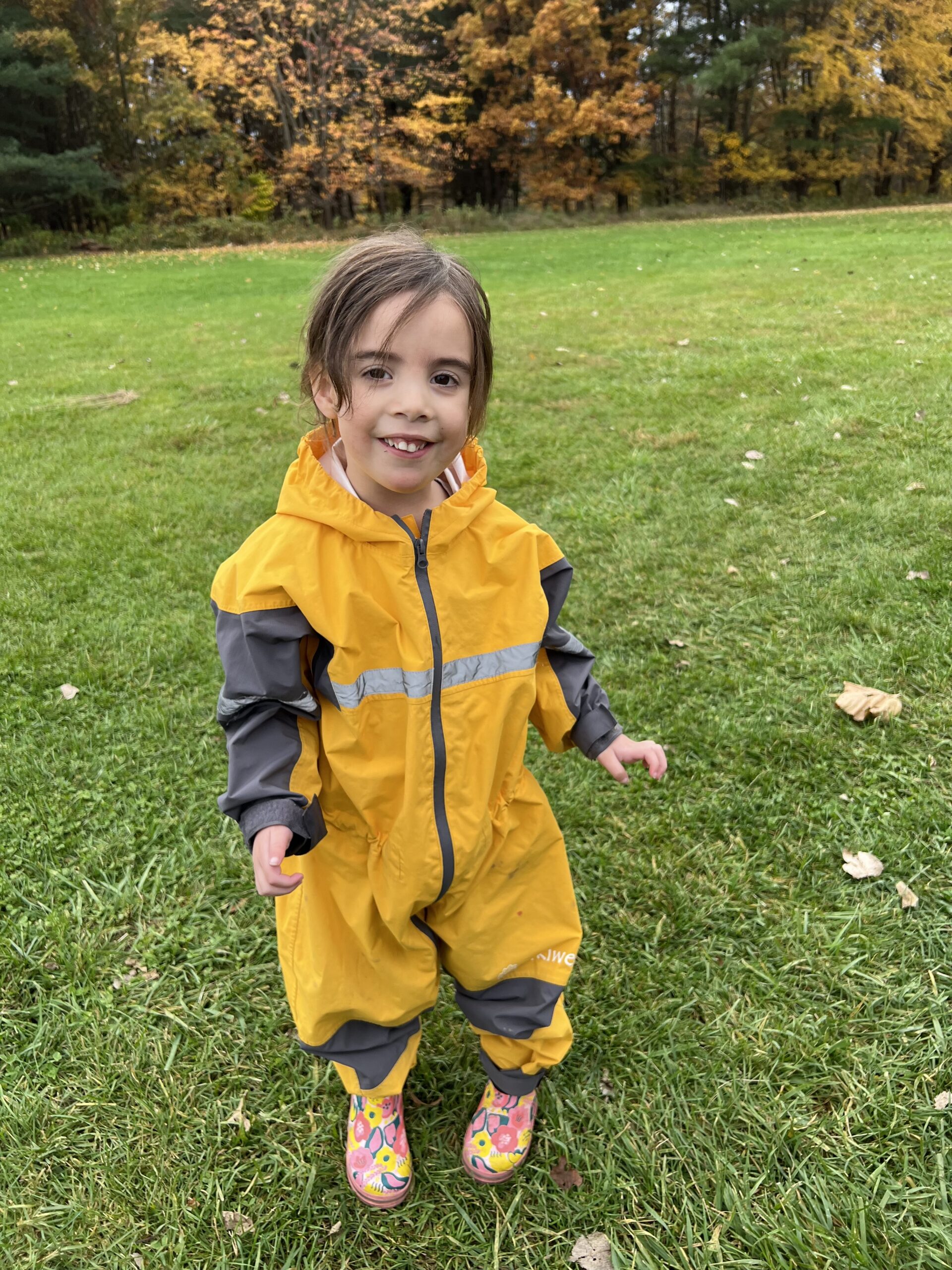 Child standing on grass wearing a yellow and black jumpsuit smiling
